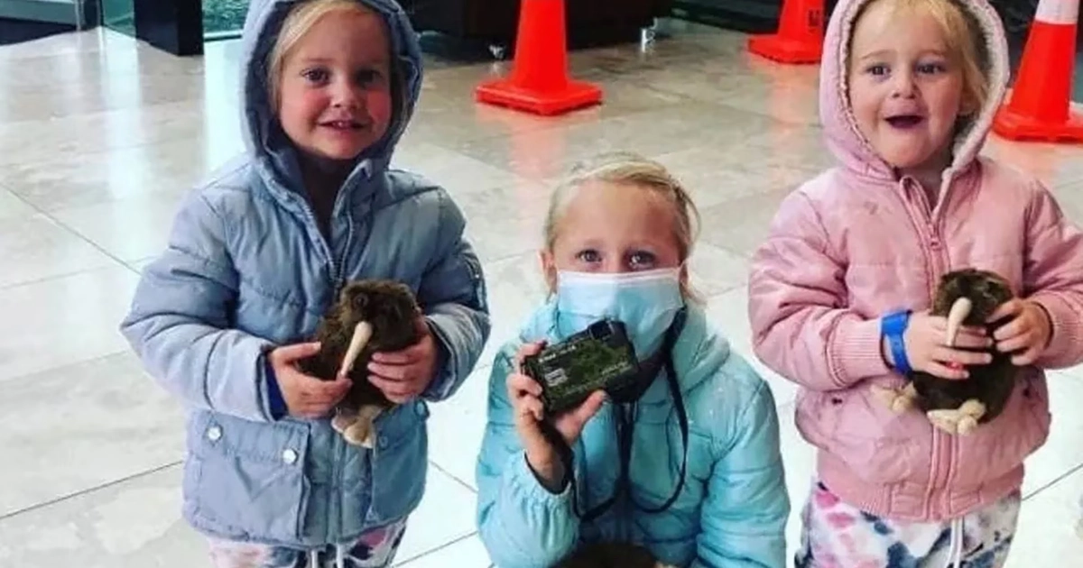 Tres niñas, dos de pie y una agachada, visten chaquetas con capucha. Dos sostienen peluches de kiwi. Una lleva mascarilla y un objeto negro. Conos naranjas al fondo