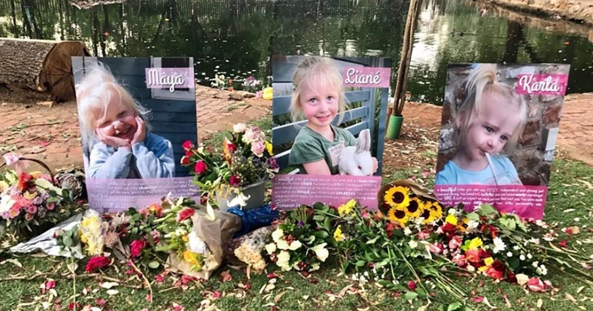Tres retratos de niñas pequeñas, Maya, Liané y Karla, se exhiben en un memorial al aire libre con flores, pétalos y un cuerpo de agua al fondo