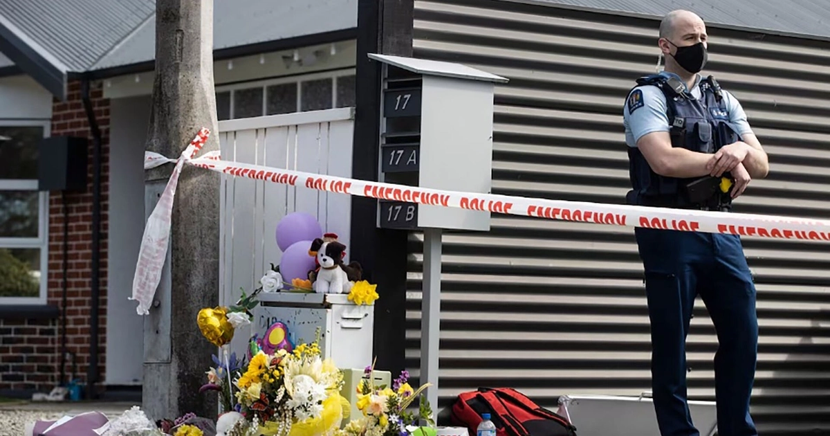 Oficial de policía con mascarilla en una calle, delante de un memorial con flores, globos y peluches, una cinta policial y buzones de correo residenciales