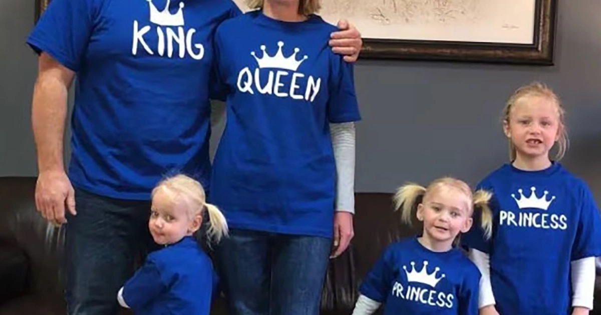Hombre, mujer y tres niñas posan; visten camisetas azules con coronas y los textos King, Queen y Princess. Detrás hay un sofá y un cuadro