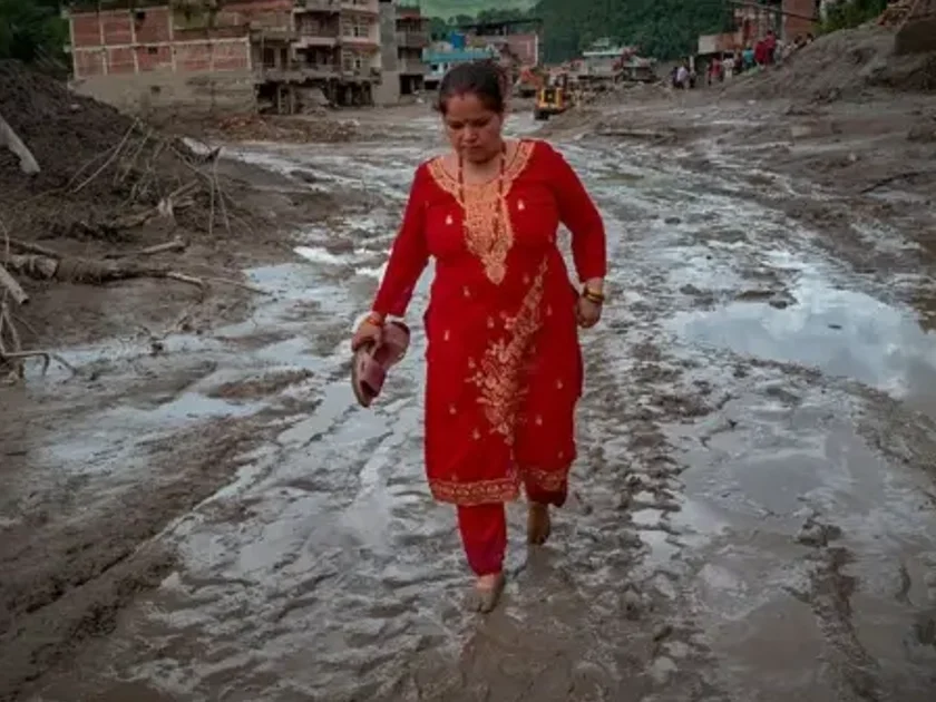 Una mujer camina descalza por un rio de lodo y detrás se ven edificios derruidos.