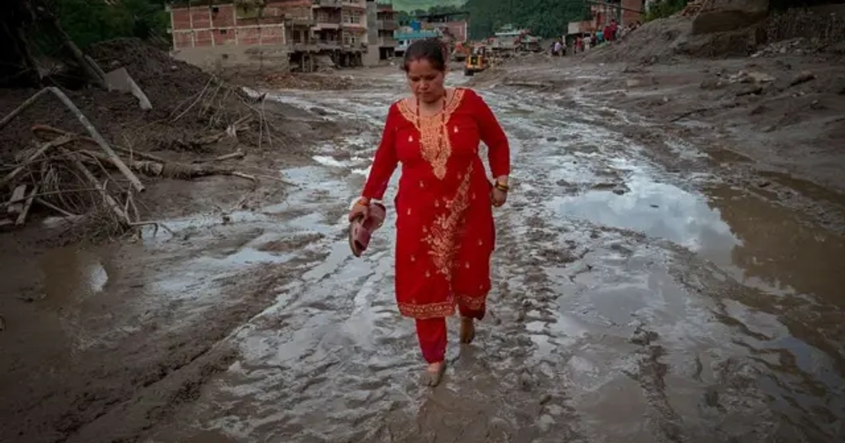 Una mujer camina descalza por un rio de lodo y detrás se ven edificios derruidos.