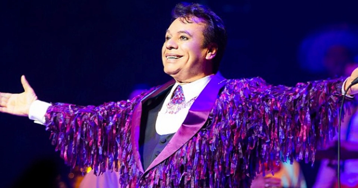 Juan Gabriel sonríe ampliamente en el escenario, vistiendo una chaqueta morada y rosa con flecos, con los brazos extendidos y un fondo oscuro
