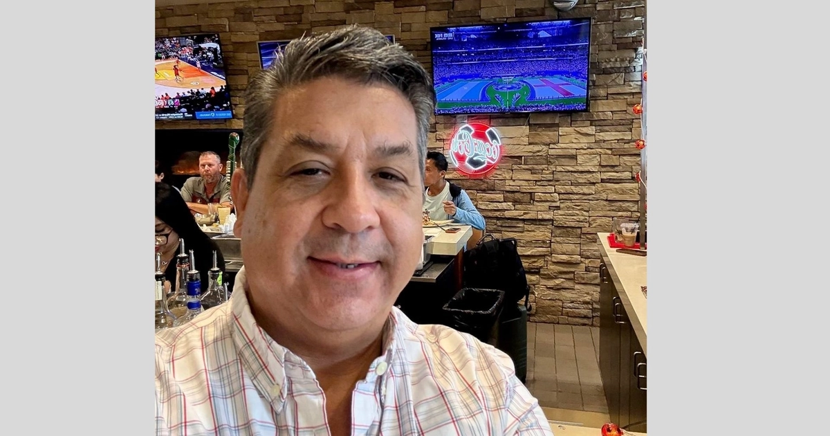 Hombre sonriente con camisa de cuadros en primer plano. Detrás, una barra de bar con botellas, televisores encendidos y una pared de piedra. Personas al fondo