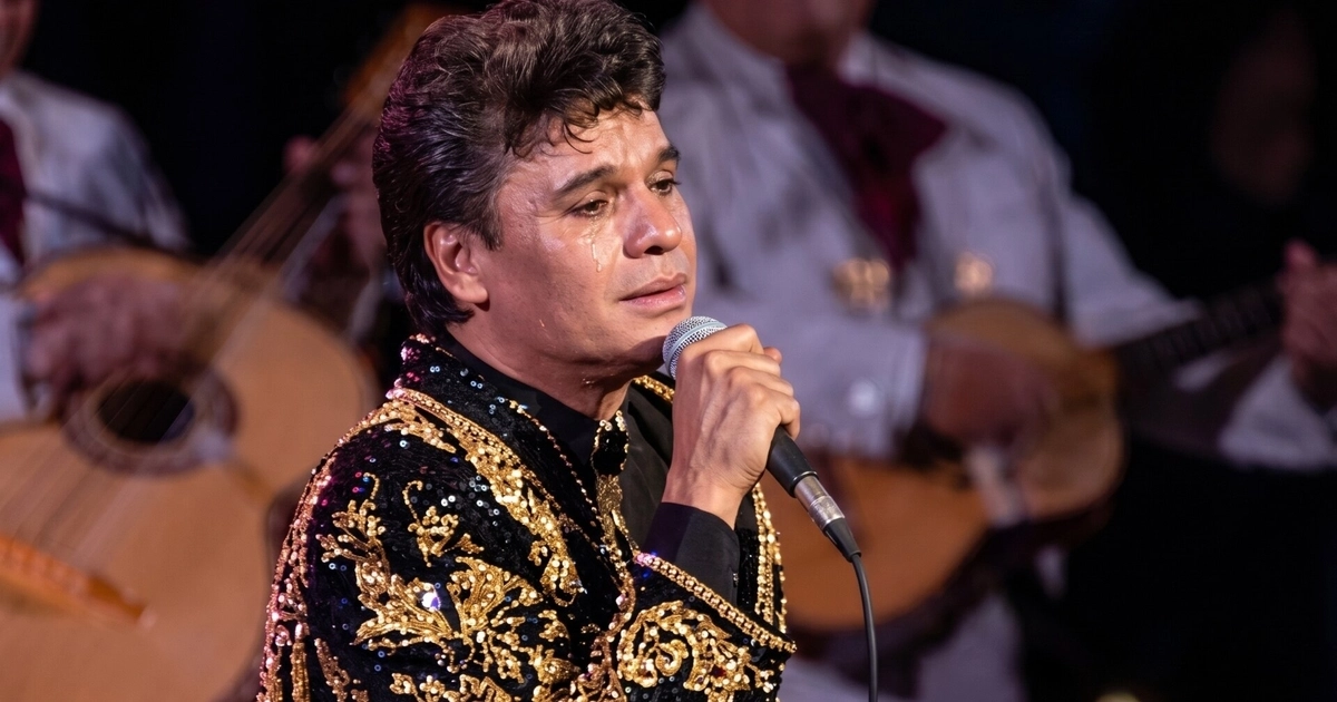 Primer plano de Juan Gabriel llorando en el escenario, vistiendo un traje negro con bordados dorados, con un micrófono en la mano y mariachis borrosos al fondo
