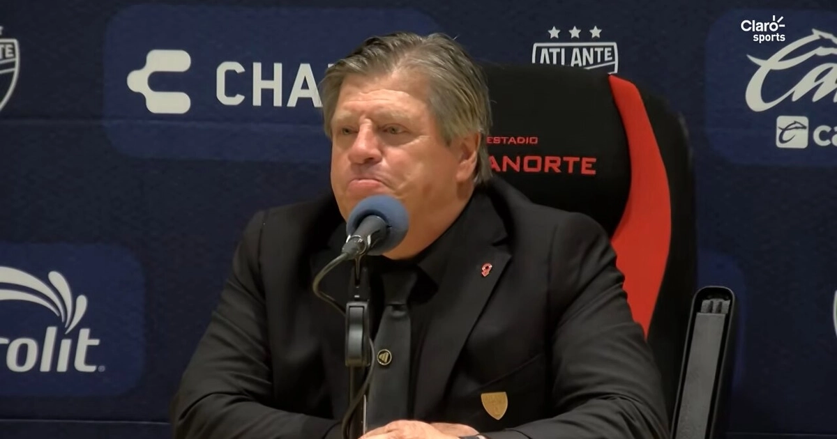 El director técnico Miguel Herrera, vestido con traje y corbata negros, permanece sentado frente a un micrófono