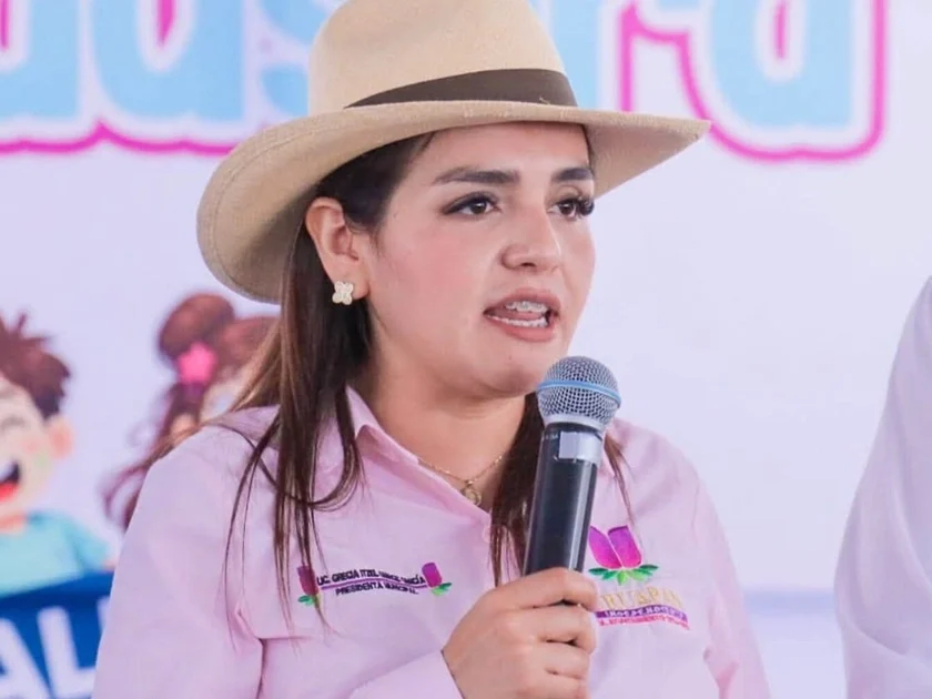 Grecia Quiroz con sombrero y camisa rosa, sosteniendo un micrófono. Se aprecian parcialmente dos hombres y un cartel con ilustraciones al fondo