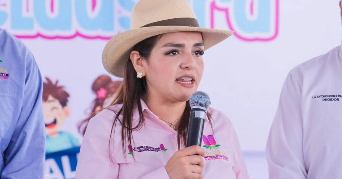 Grecia Quiroz con sombrero y camisa rosa, sosteniendo un micrófono. Se aprecian parcialmente dos hombres y un cartel con ilustraciones al fondo