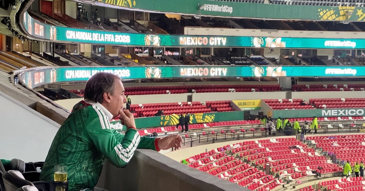 Hombre sentado de perfil con camiseta verde en un palco de estadio. En las pantallas se lee 'Copa Mundial de la FIFA 2026' y 'Mexico City'.