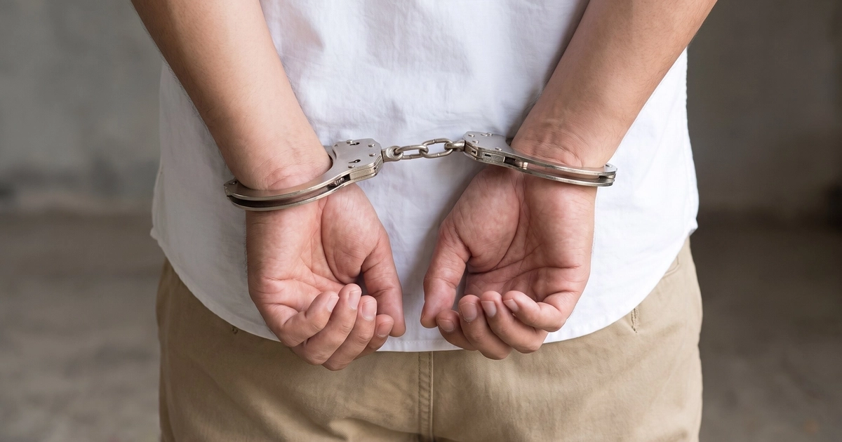 Un joven viste una camisa blanca y un pantalón beige, mientras sus manos están esposadas por detrás de su espalda. (Imagen Ilustrativa Infobae)