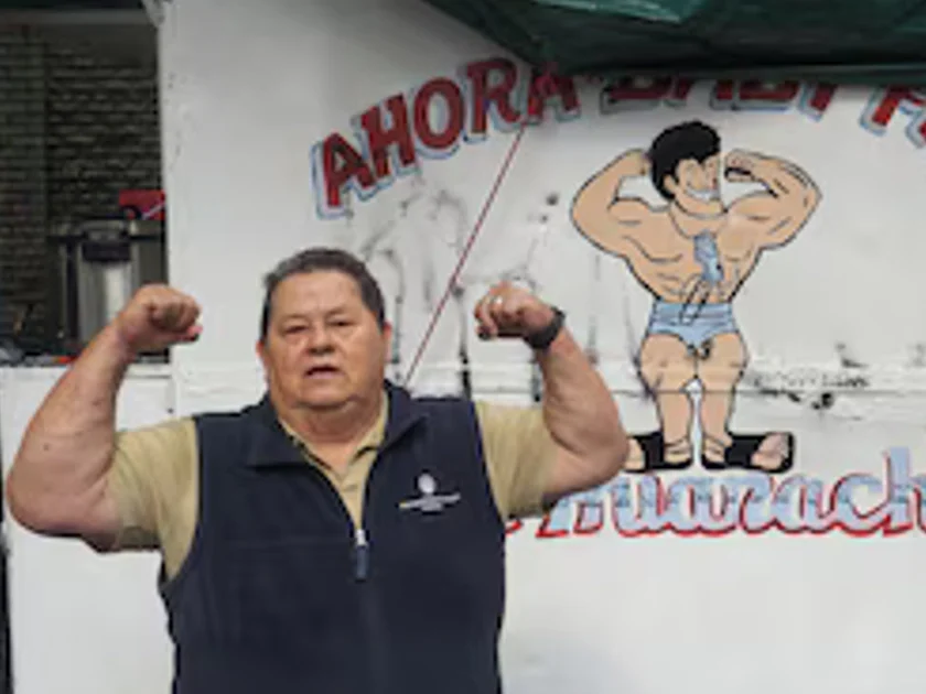 Un hombre mayor flexiona sus bíceps frente a un puesto blanco con la ilustración de un hombre musculoso y el letrero Huaraches.