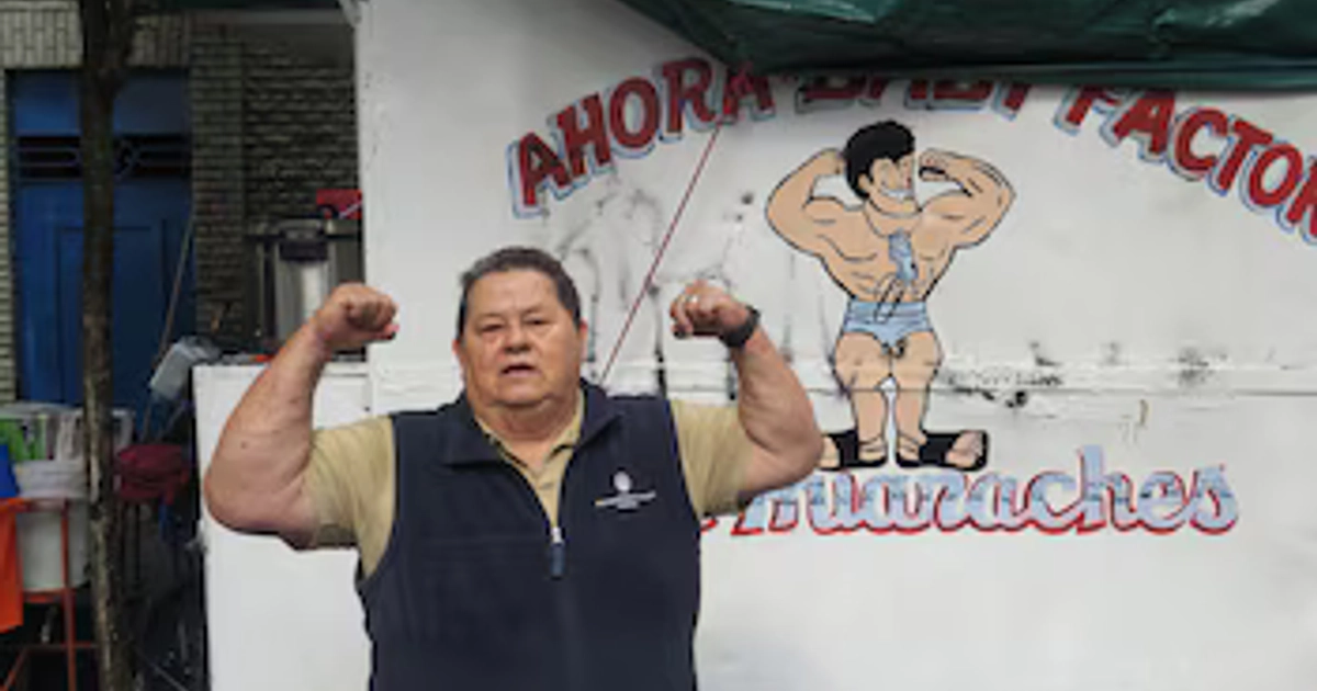 Un hombre mayor flexiona sus bíceps frente a un puesto blanco con la ilustración de un hombre musculoso y el letrero Huaraches.