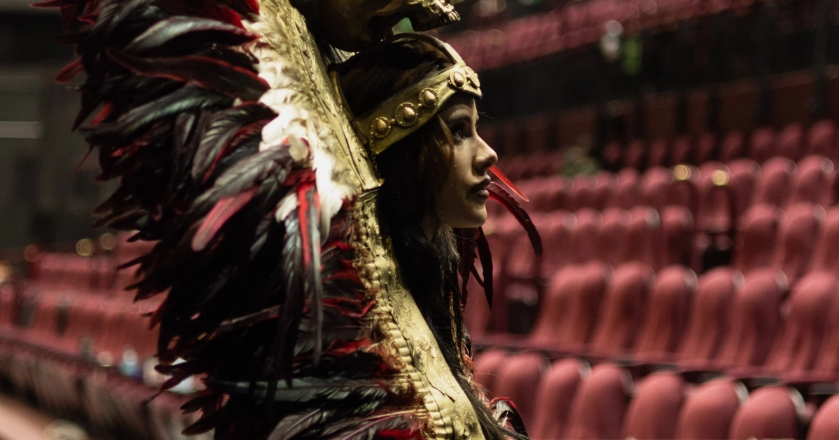 La actriz Gala Montes posa de perfil con un atuendo ceremonial de plumas y un adorno de calavera en el área de butacas de un teatro. (@malinchethemusical/Instagram)