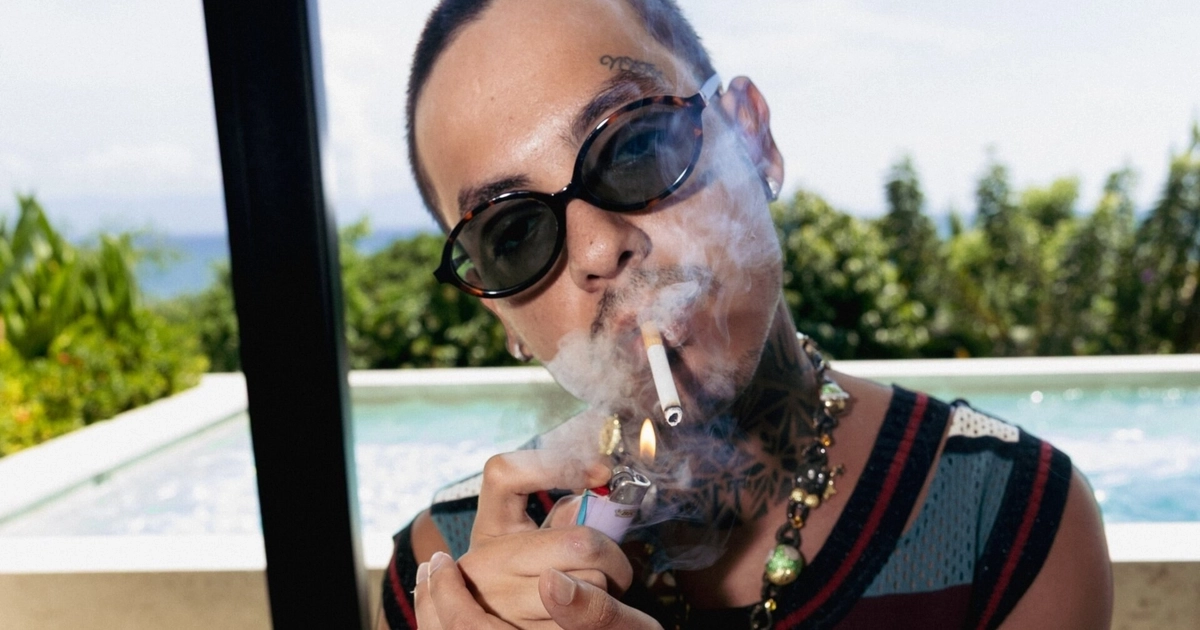 Natanael Cano lleva gafas de sol y enciende un cigarrillo con un encendedor, frente a una piscina