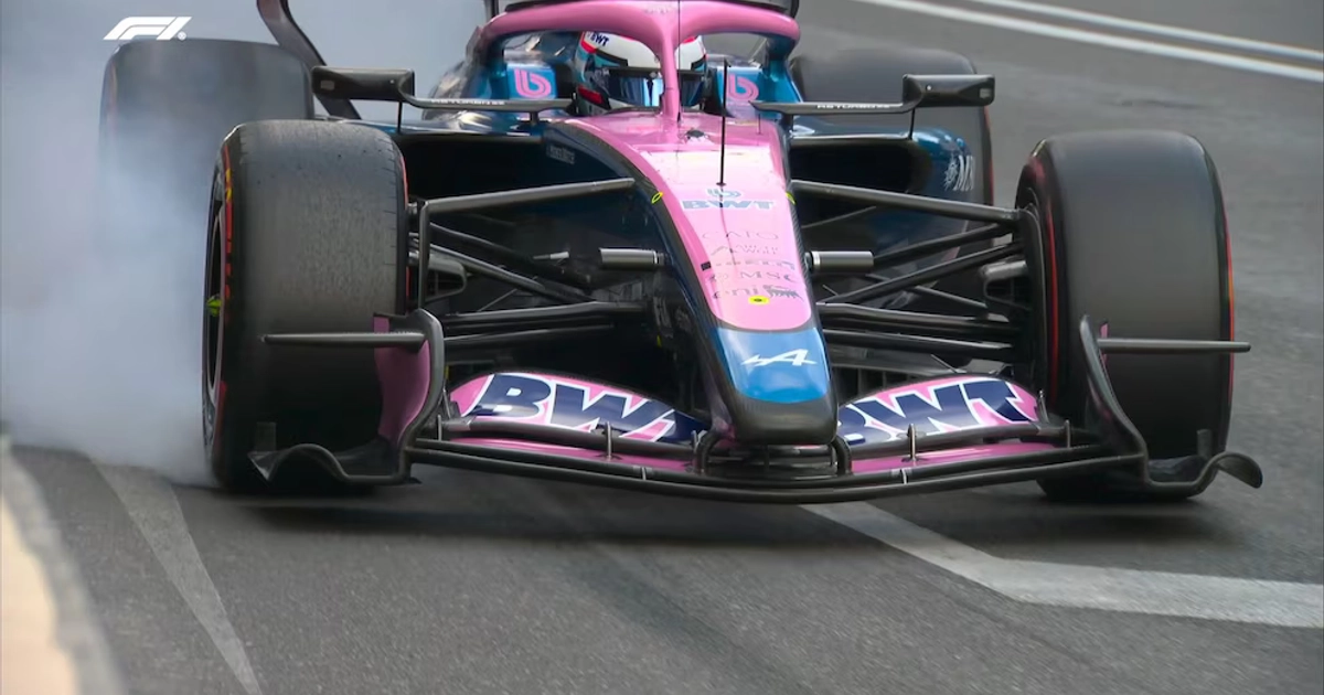 Monoplaza de carreras rosa y azul sobre la pista con humo saliendo de uno de sus neumáticos delanteros por el frenado