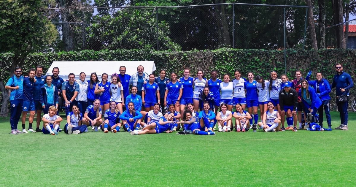 Un grupo grande de mujeres y hombres con uniformes deportivos azules y blancos posa en un campo de césped verde. Hay una valla con vegetación al fondo.