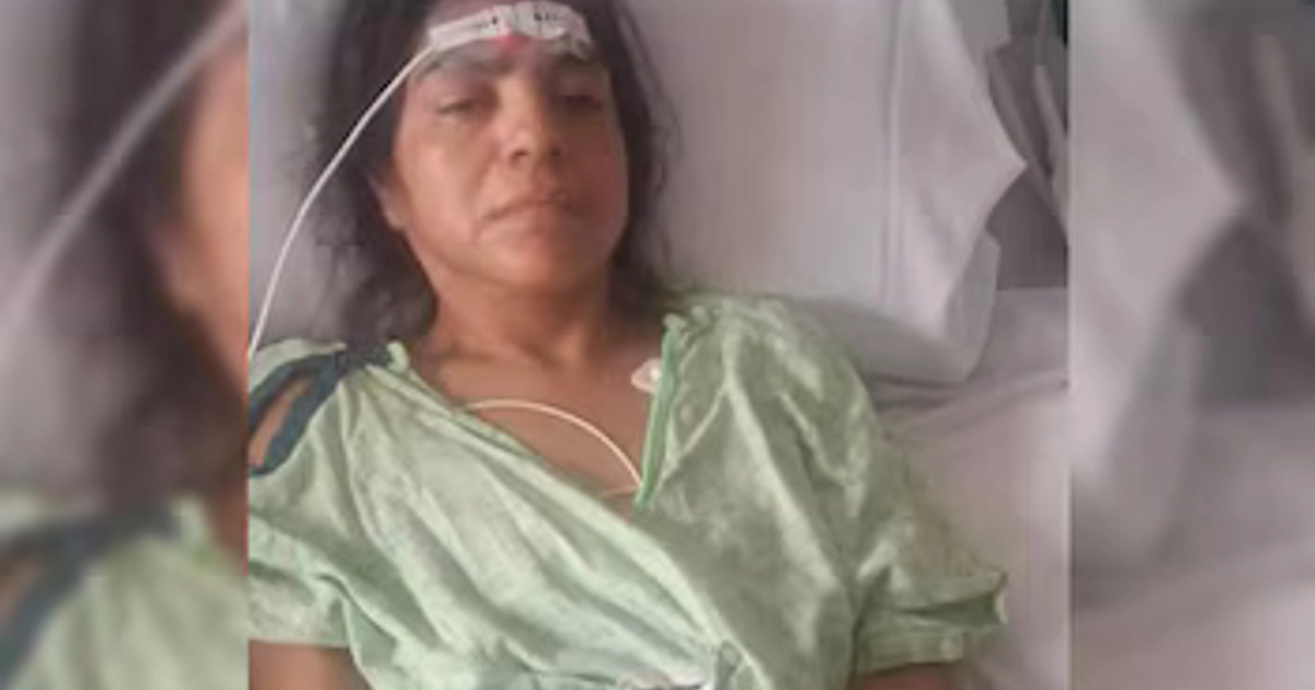 Una mujer recostada en una cama de hospital lleva una bata verde y un sensor en la frente