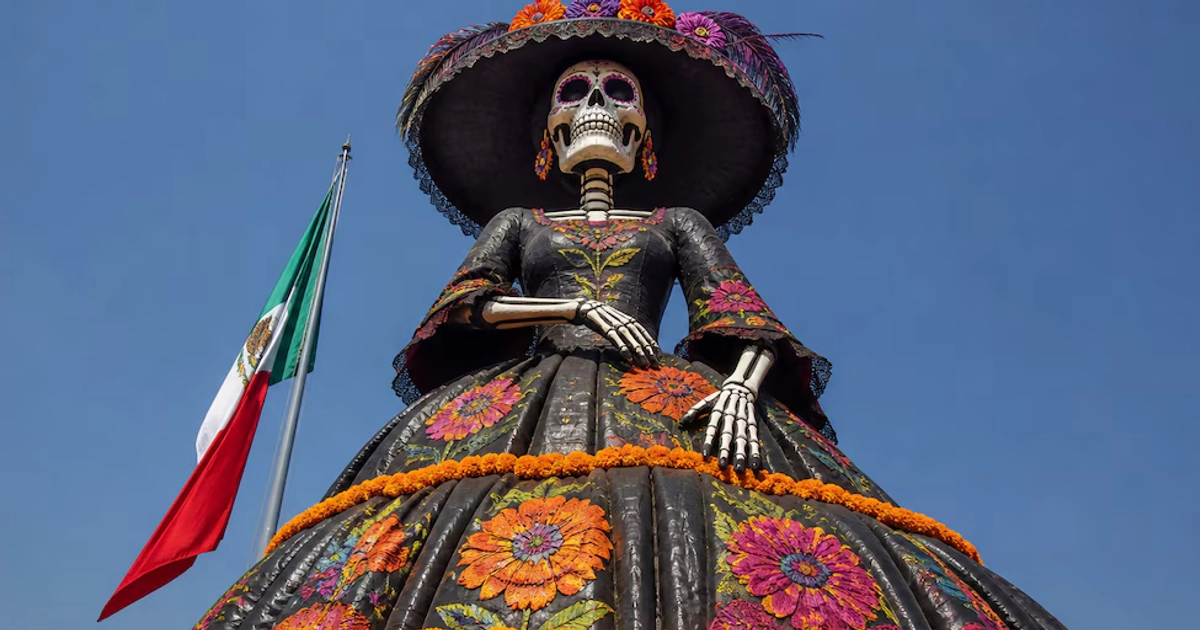 Figura de una catrina con sombrero y vestido oscuro rodeada de flores de cempasúchil, con la bandera de México y el cielo de fondo.
