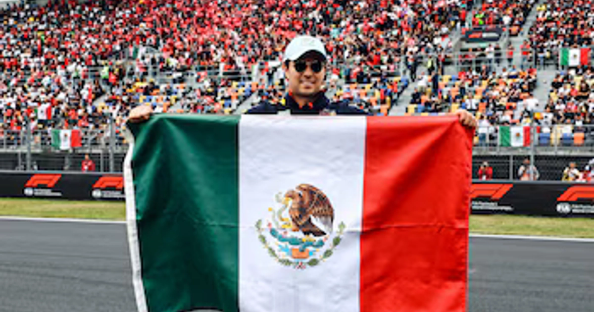 Un hombre con gorra y gafas oscuras sostiene la bandera de México desplegada en una pista frente a gradas con público.
