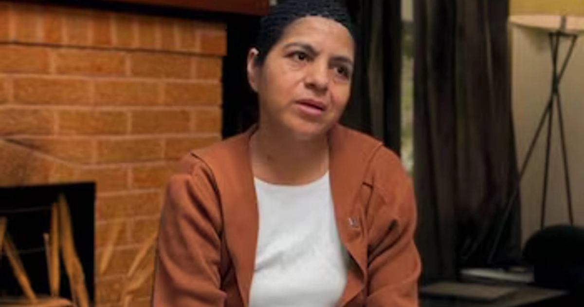 Una mujer con gorro negro y chaqueta marrón permanece sentada ante una chimenea de ladrillo