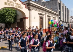 Estudiantes con uniforme escolar salen por el arco de la Escuela Primaria Benito Juárez junto a adultos y un vendedor de globos.
