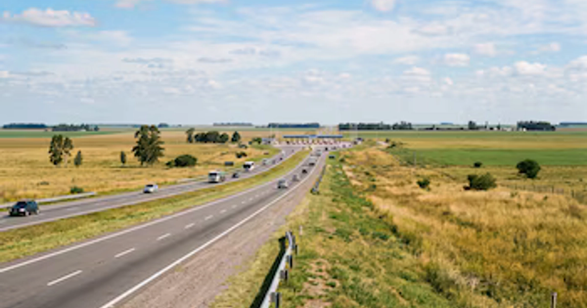 Una autopista con automóviles y camiones atraviesa un área rural con vegetación y cabinas de peaje al fondo bajo un cielo azul