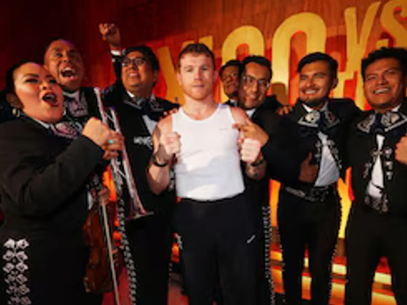 Saúl Canelo Álvarez en camiseta blanca junto a varios músicos de mariachi con trajes negros e instrumentos musicales frente a un panel.