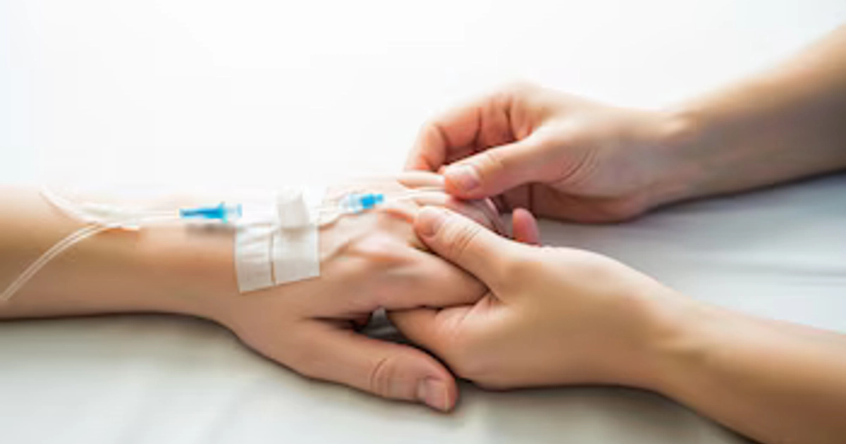 Dos manos entrelazadas sobre una sábana blanca; una mano con catéter intravenoso y adhesivo médico, la otra mano la sujeta.