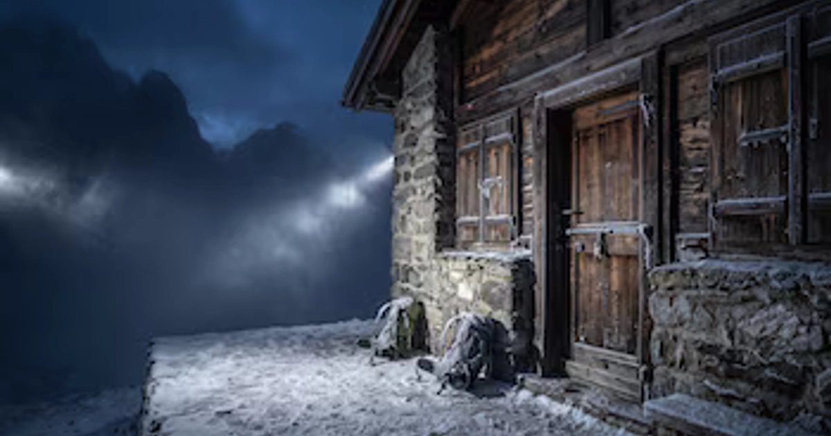 Fachada de un refugio de piedra y madera cerrado con candado, dos mochilas cubiertas de nieve en el suelo y montañas de fondo de noche.