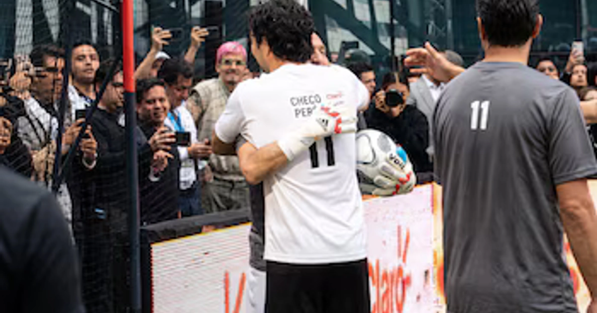 Sergio Pérez de espaldas con guantes y un balón de fútbol abraza a un hombre frente a varias personas que toman fotos con sus teléfonos.